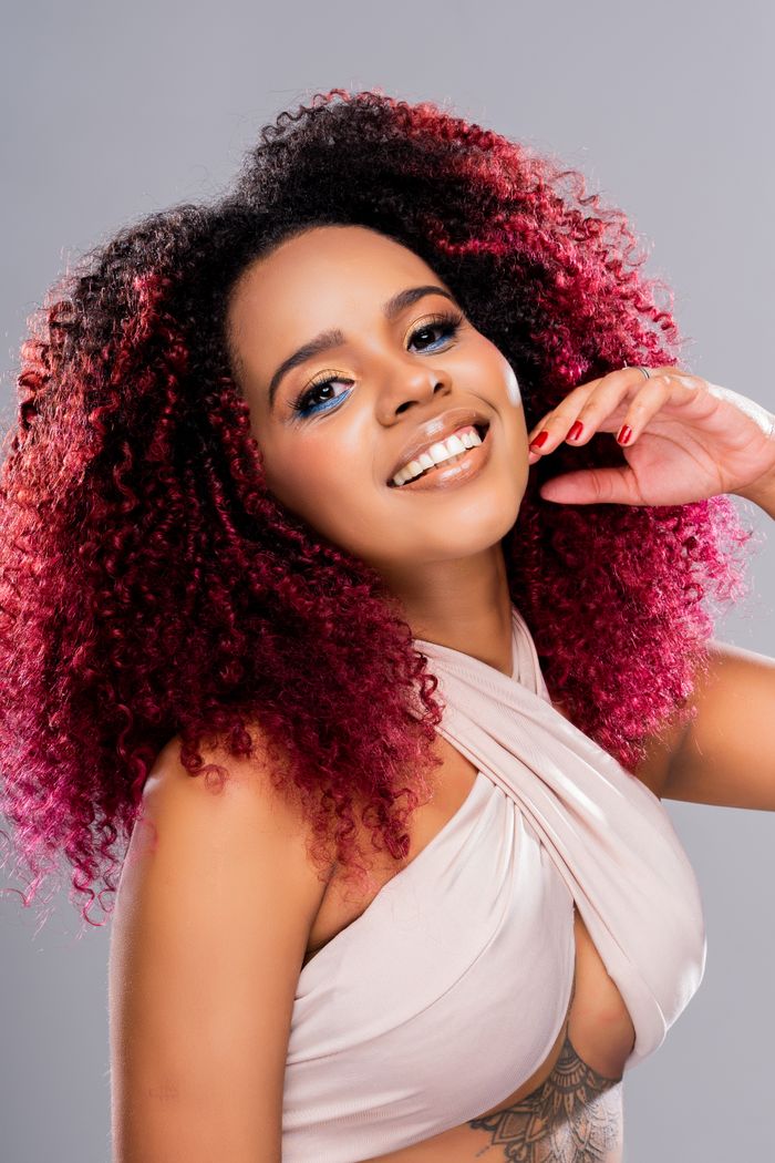 Studio portrait of a smiling woman with curly dyed red hair, posing against a gray background.