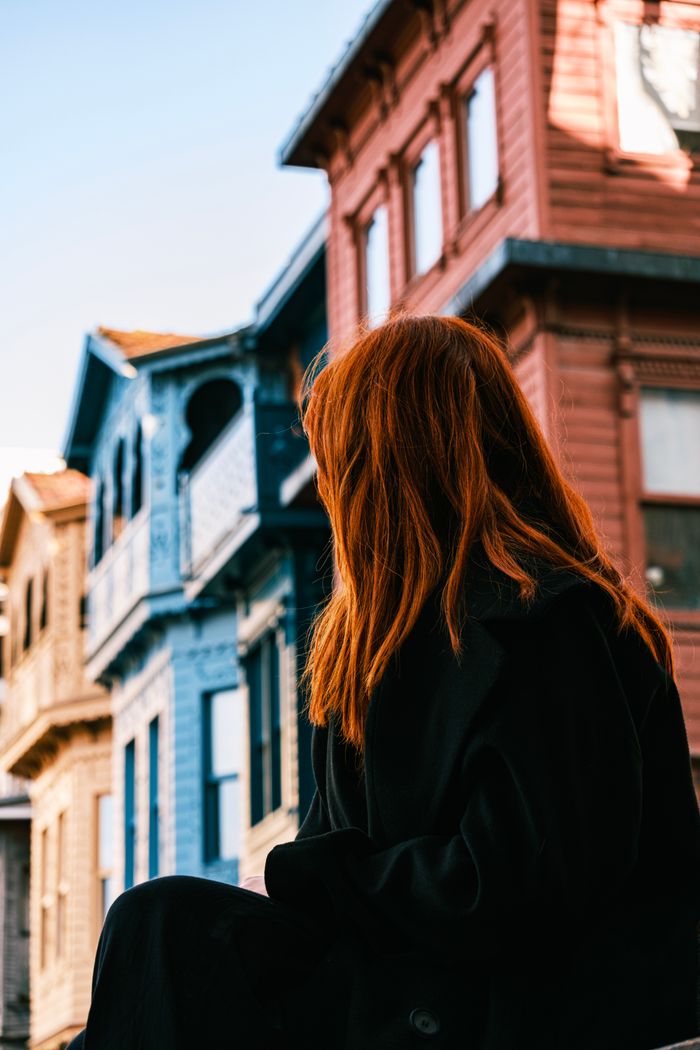 A red-haired woman sitting in an urban area with colorful historical buildings.